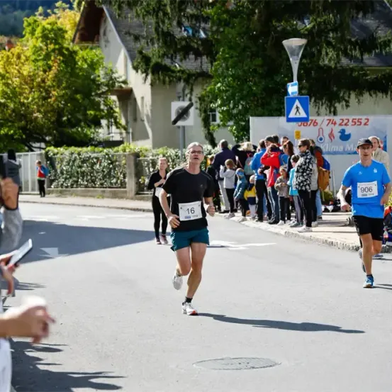Ein Marathonläufer mit der Nummer 16 läuft auf der Straße, gefolgt von einem weiteren Läufer mit der Nummer 906. Zuschauer beobachten sie.