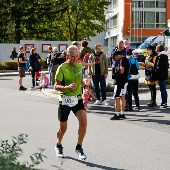 Ein Mann in einem grünen Shirt und schwarzen Shorts läuft auf einer Straße mit einer Nummer 904. Mehrere Leute schauen zu.