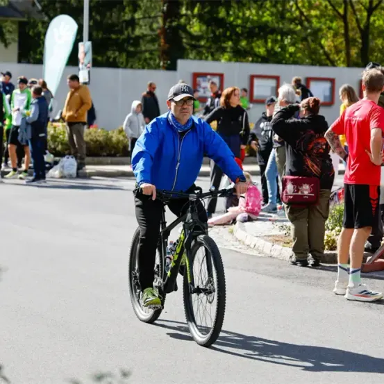Ein Mann fährt mit dem Fahrrad eine Straße hinunter, während eine Menschenmenge am Straßenrand zusieht. Bäume und Banner säumen die Straße.