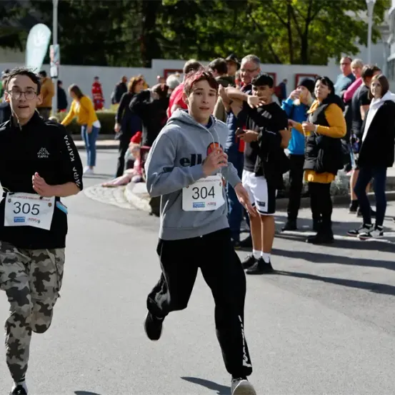 Zwei Läufer nehmen an einem Rennen teil, einer trägt eine graue Kapuzenjacke und der andere ein schwarzes Sweatshirt. Zuschauer schauen zu, einige stehend und andere sitzend. Bäume und Gebäude säumen die Straße.