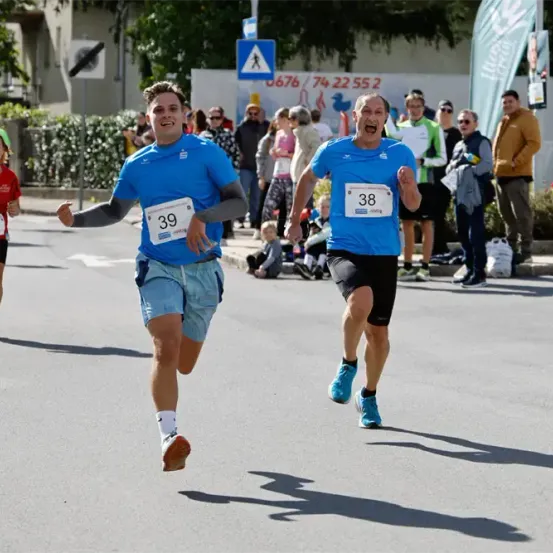 Zwei Männer in blauen Shirts laufen einen Marathon, Läufer 39 führt. Zuschauer beobachten von der Seite, einer hält ein Banner.