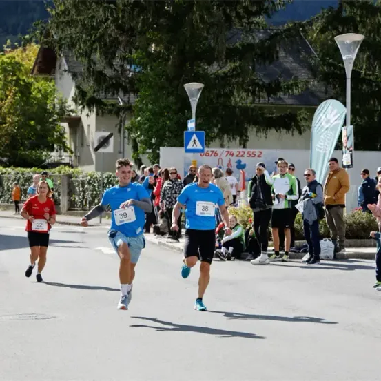 Zwei Männer mit blauen Shirts und den Nummern 39 und 38 laufen in einem Marathon, gefolgt von einer Frau mit einem roten Shirt und der Nummer 22. Zuschauer stehen am Straßenrand.