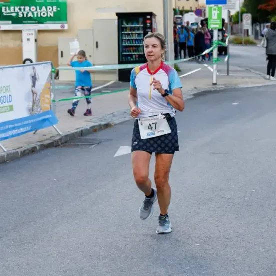 Eine Frau mit der Nummer 47 auf ihrem Shirt läuft auf der Straße einen Marathon. Im Hintergrund stehen Menschen hinter einer Seilbarriere.