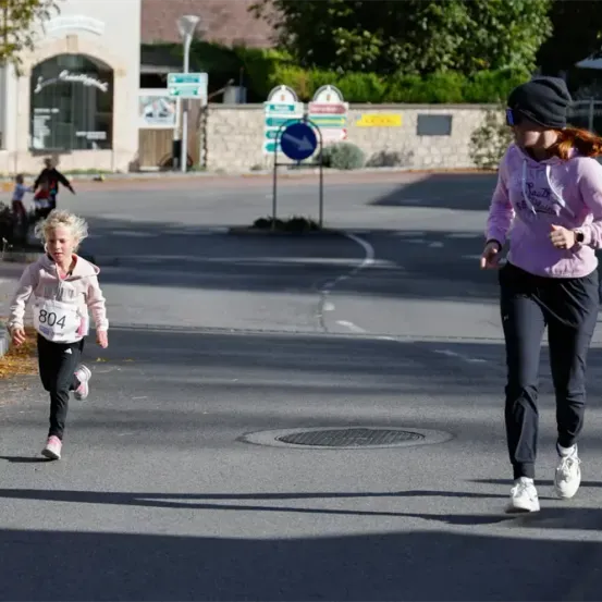 Ein Mädchen mit der Nummer 804 läuft auf einer Straße, während eine Frau in der Nähe geht, beide tragen rosa Sweatshirts und schwarze Hosen. Dahinter stehen Bäume, Pflanzen und Gebäude.