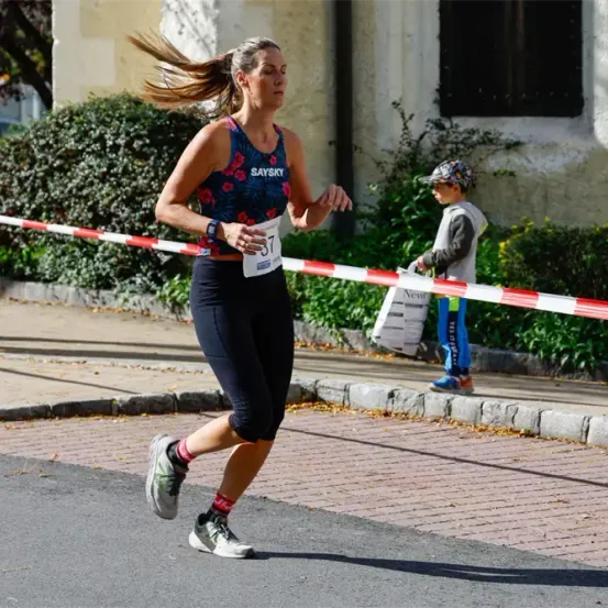 Eine Frau in einer Laufbekleidung mit der Nummer 37 läuft bei einem Marathon auf einer Stadtstraße. Hinter ihr steht ein Junge und hält eine Tasche.