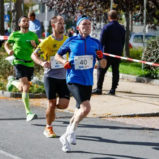 Drei Läufer nehmen an einem Marathon teil. Der Mann vorne trägt ein blaues Shirt mit der Nummer 40. Dahinter läuft ein Mann in einem gelben Shirt. Ein weiterer Mann in einem grünen Shirt folgt dahinter.