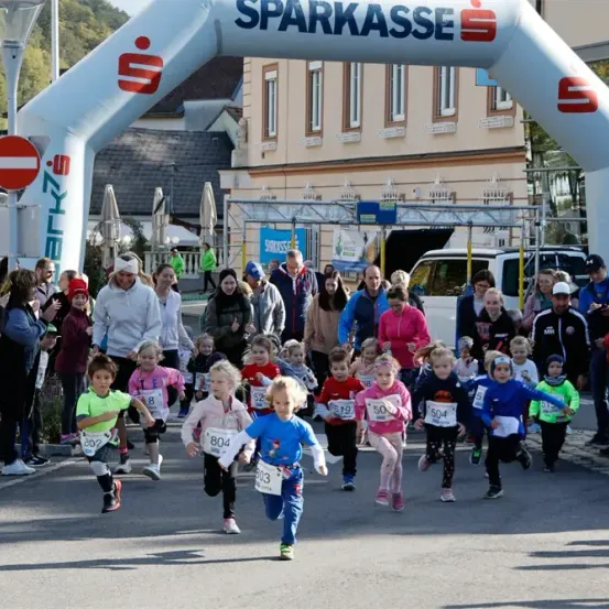 Eine Gruppe von Kindern nimmt an einem Laufwettbewerb teil, mit Nummern auf ihren Trikots, unter einem weißen Bogen mit der Aufschrift 'Sparkasse', mit Zuschauern an der Seite und einem Gebäude im Hintergrund.
