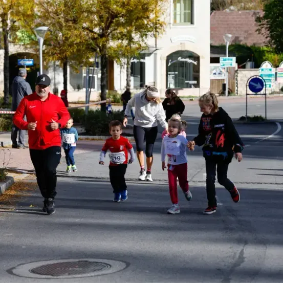 Eine Gruppe von Menschen läuft bei einem Marathon. Ein erwachsener Mann führt, gefolgt von mehreren Kindern mit Nummern auf ihren Trikots. Sie laufen auf der Straße und passieren Bäume und Gebäude.