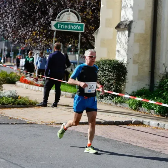 Ein Mann läuft bei einem Marathon auf einer Straße mit einem Schild, das 'Friedhofe' steht. Er trägt eine Sonnenbrille und ein Shirt mit der Nummer 28. Hinter ihm steht ein Mann in der Nähe einer Absperrung.
