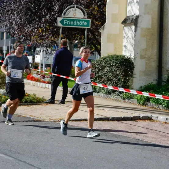 Zwei Läufer nehmen an einem Marathon teil, einer mit der Nummer 21 und der andere mit 47. Sie laufen auf einer Straße mit einer Absperrung und einem Schild, das 'Friedhofe' steht.