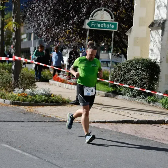 Ein Läufer mit einem grünen Shirt mit der Nummer 314 nimmt an einem Marathon teil und läuft an einem Friedhof-Schild vorbei.