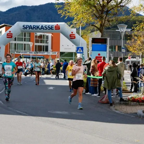 Läufer nehmen vor einem Sparkasse-Gebäude mit Bergen im Hintergrund an einem Marathon teil.