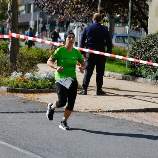 Eine Frau in einem grünen Shirt läuft auf der Straße, während ein Mann sie vom Bürgersteig aus beobachtet. Bäume und eine rot-weiße Barriere sind im Hintergrund.