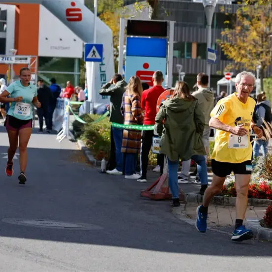 Eine Frau mit der Nummer 307 und ein Mann mit der Nummer 8 laufen auf einer Straße, während mehrere Menschen zuschauen. Die Straße ist mit Straßenschildern und Absperrungen versehen. Im Hintergrund befindet sich ein Gebäude mit Glasfenstern.