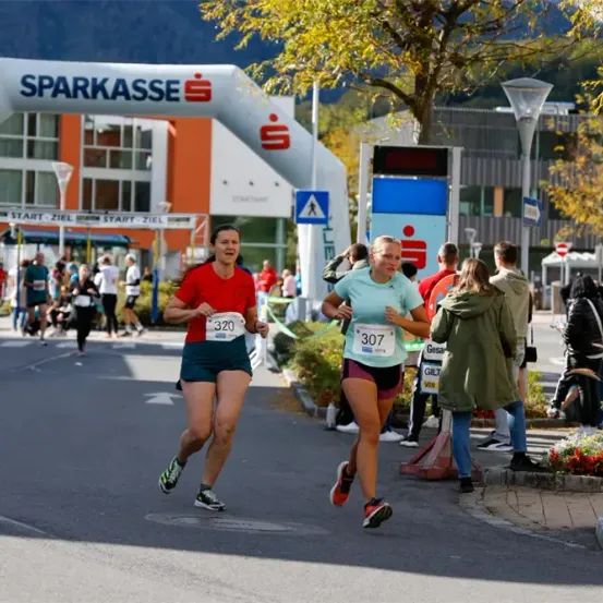 Zwei Läuferinnen treten bei einem von Sparkasse gesponserten Marathon an, mit Zuschauern und Bäumen im Hintergrund.