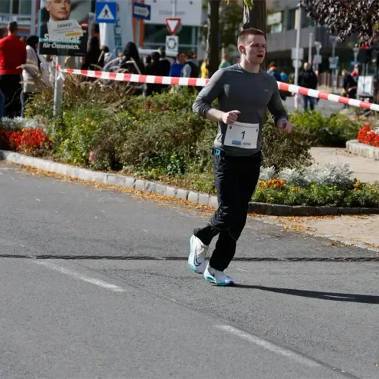 Ein Läufer mit der Nummer 1 auf dem Trikot nimmt an einem Marathon auf einer Stadtstraße teil, mit Zuschauern und Absperrungen in der Nähe.