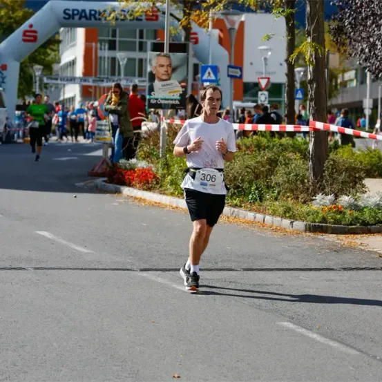 Ein Läufer mit der Startnummer 306 läuft auf einer Straße mit anderen Läufern, einem weißen Auto und einem weißen Bogen mit dem Text Spartakus.