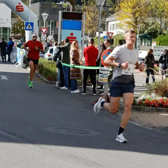 Läufer nehmen an einem Marathon teil, wobei ein Mann in einem roten Shirt und ein anderer in einem weißen Shirt die Führung übernimmt. Zuschauer säumen die Straße, und Straßenschilder und ein Banner sind sichtbar.