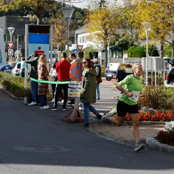 Eine Marathonläuferin in einem grünen Shirt und schwarzen Shorts läuft eine Straße entlang, an einem Banner mit der Aufschrift 'Gesantmer P'. Zuschauer beobachten das Ereignis.