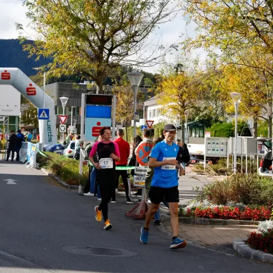 Ein Marathon ist unterwegs, mit Läufern auf der Straße, umgeben von Bäumen und Gebäuden. Eine Frau mit Startnummer 17 und ein Mann in Blau laufen.