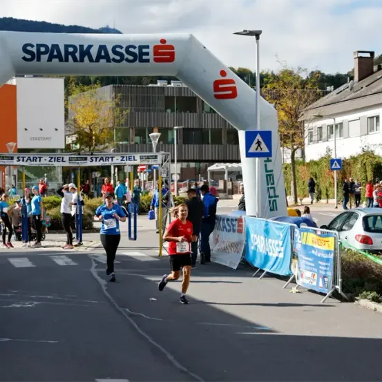Ein Marathon ist im Gange, mit Läufern, die unter einem weißen Bogen mit dem Wort 'Sparkasse' darauf passieren. Zuschauer beobachten von den Seitenlinien.