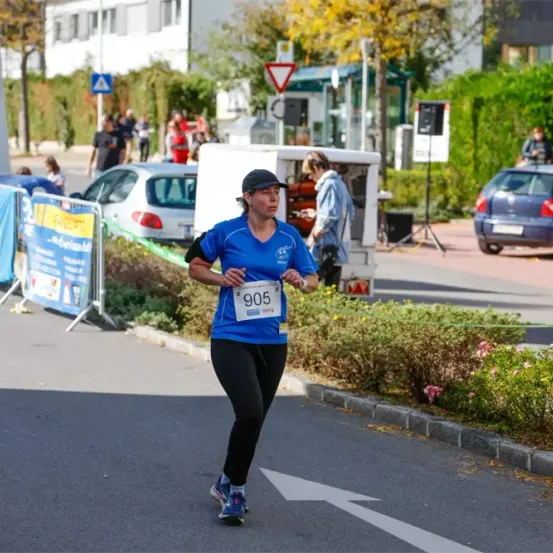 Eine Marathonläuferin mit der Nummer 905 läuft auf der Straße. Es gibt geparkte Autos, Pflanzen und Gebäude an den Seiten.