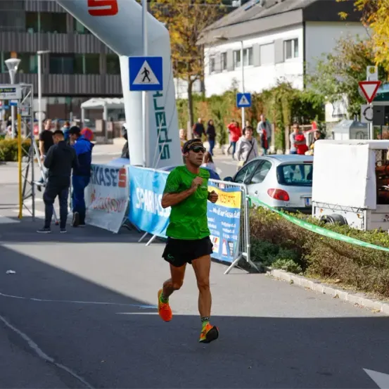 Ein Läufer in einem grünen Shirt und schwarzen Shorts nimmt an einem Marathon teil. Der Läufer läuft an einem Gebäude und Bäumen vorbei. Mehrere Menschen stehen auf dem Bürgersteig.