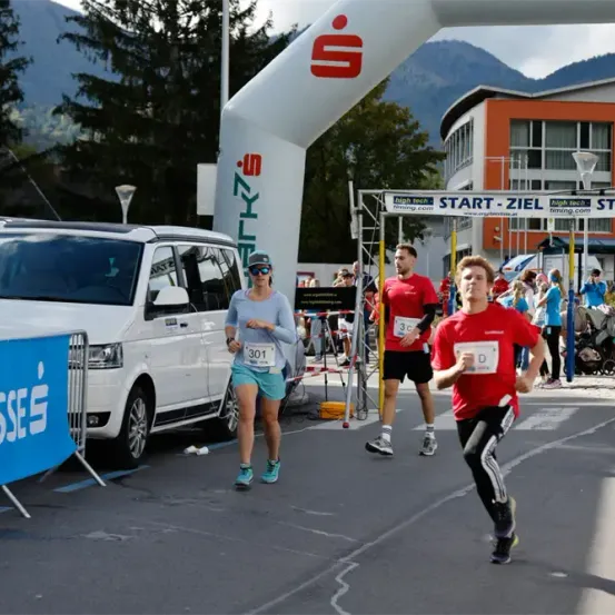 Läufer mit Nummern nehmen an einem Marathon teil und passieren ein Schild mit der Aufschrift 'Start-Ziel'. Dahinter ist ein weißer Van und ein Banner zu sehen.