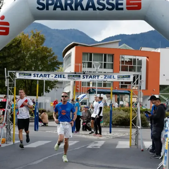 Ein Marathon ist im Gange, mit einem Mann in Blau, der führt, gefolgt von einem Läufer in Weiß. Zuschauer beobachten von den Seitenlinien, und ein Gebäude mit orangefarbenen Wänden ist im Hintergrund sichtbar.
