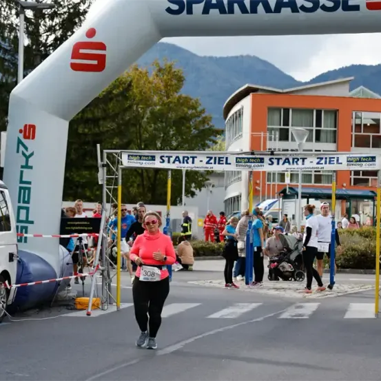 Eine Läuferin mit der Nummer 308 läuft einen Marathon und passiert einen weißen Bogen. Es stehen Leute herum und beobachten das Rennen. Im Hintergrund befinden sich Gebäude und Berge.