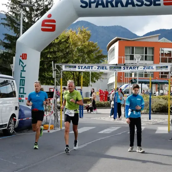 Drei Läufer in verschiedenen Farben laufen auf der Straße unter einem weißen Bogen mit der Aufschrift 'Sparkasse'.