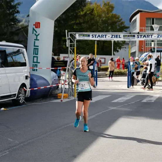 Eine Marathonläuferin mit der Nummer 31 auf ihrem Shirt läuft auf einer Stadtstraße. Hinter ihr ist ein Banner mit dem Text START-ZIEL. Auf der linken Seite steht ein Van. In der Ferne stehen Menschen, von denen einige stehen.