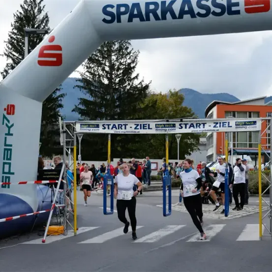 Ein Marathon ist im Gange, Teilnehmer laufen unter einem Start-Ziel-Bogen. Zuschauer und Offizielle sind an den Seitenlinien anwesend.
