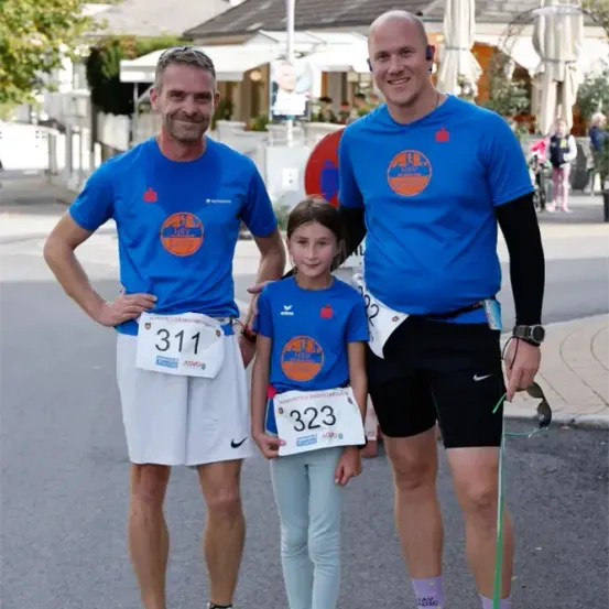 Drei Personen stehen auf einer Straße und tragen passende blaue T-Shirts mit weißen Nummern und weißen Shorts. Der Mann auf der linken Seite trägt eine Sonnenbrille, während der Mann auf der rechten Seite eine Leine hält. Sie werden von einem jungen Mädchen mit der Nummer 323 auf dem Shirt begleitet. Hinter ihnen befinden sich Gebäude, Bäume und Menschen.