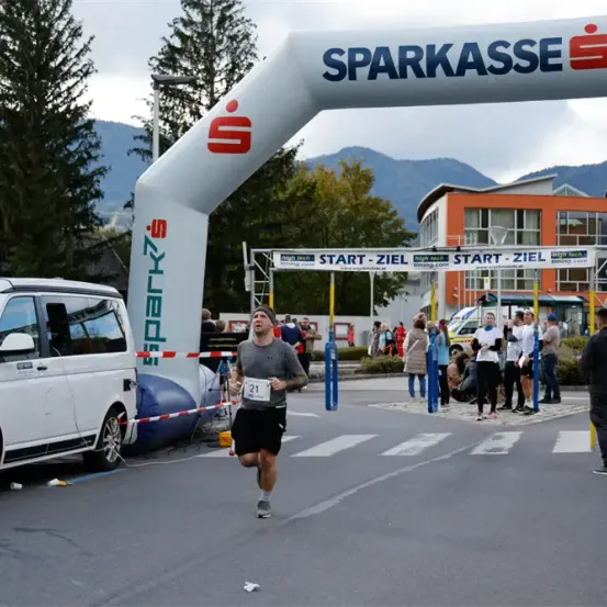 Ein Marathonläufer läuft unter einem Torbogen mit der Aufschrift Sparkasse vorbei, während mehrere Zuschauer zuschauen. Im Hintergrund befinden sich Berge und ein Gebäude mit orangefarbenen Wänden.
