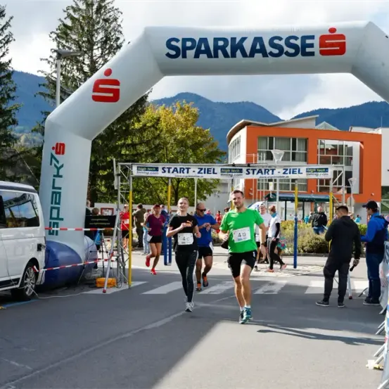 Ein Marathonlauf findet statt, bei dem Teilnehmer unter einem weißen Bogen mit der Aufschrift 'Sparkasse' laufen. Das Rennen hat eine weiße Ziellinie mit gelben Stangen. Im Hintergrund sind Bäume und Berge zu sehen.