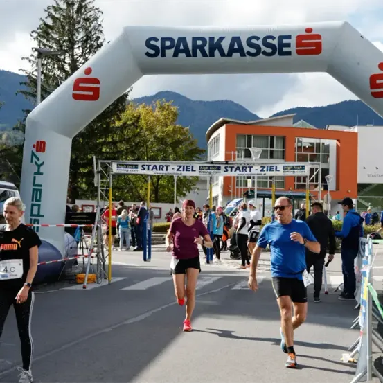 Läufer laufen unter einem weißen Bogen mit dem Text Sparkasse oben. Im Hintergrund sind Gebäude, Bäume und Berge zu sehen.