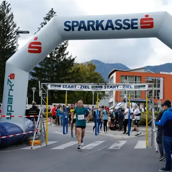 Ein Läufer durchquert ein Zielbogentor, mit Zuschauern und Bergkulisse im Hintergrund. Der Bogen trägt die Aufschrift 'SPARKASSE'.