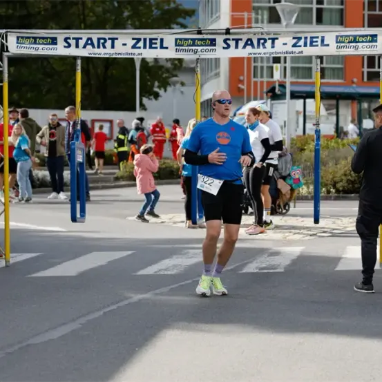 Ein Marathonläufer mit der Nummer 322 läuft unter einem Schild vorbei, das START-ZIEL sagt. Hinter ihm stehen und beobachten Leute, einschließlich eines Mannes mit einer Jacke.