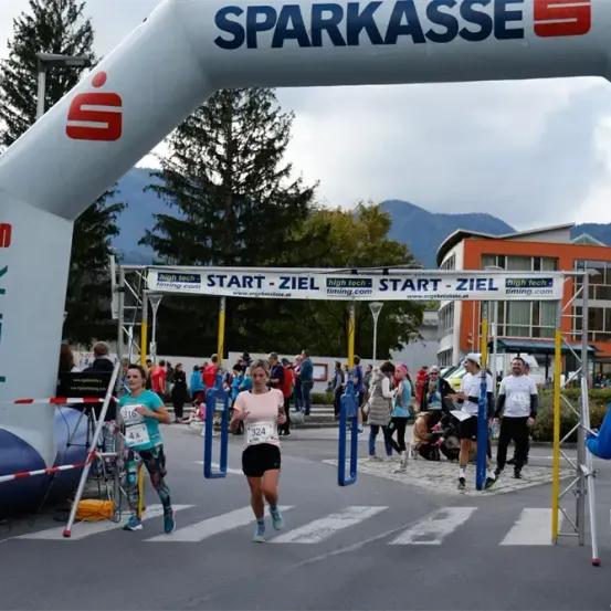 Läufer überqueren die Ziellinie eines Marathons. Ein Bogen trägt die Aufschrift Sparkasse und Start-Ziel. Eine Menge schaut zu, wie Läufer die Ziellinie überqueren.