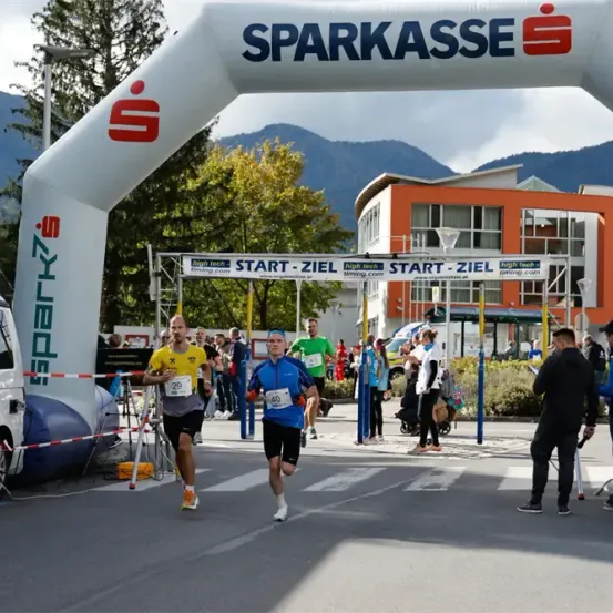 Ein Marathon-Event findet statt, bei dem Läufer durch einen großen aufblasbaren Bogen gehen. Zuschauer stehen am Straßenrand, und einige halten die Momente mit Kameras fest. Ein Van ist in der Nähe geparkt.