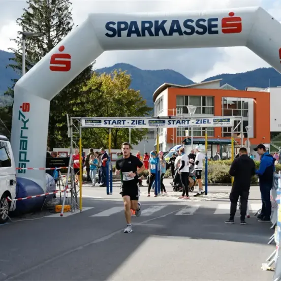 Ein Läufer überquert die Ziellinie eines Marathons unter einem weißen Bogen, gesponsert von 'Sparkasse'. Zuschauer stehen am Straßenrand. Im Hintergrund befinden sich Gebäude und Berge.