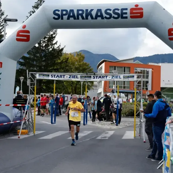 Ein Marathonläufer läuft unter einem weißen Bogen mit der Aufschrift 'SPARKASSE' hindurch. Zuschauer stehen an der Straße, einige tragen Hüte und Turnschuhe. Ein blau-weißes aufblasbares Bauwerk befindet sich in der Nähe.