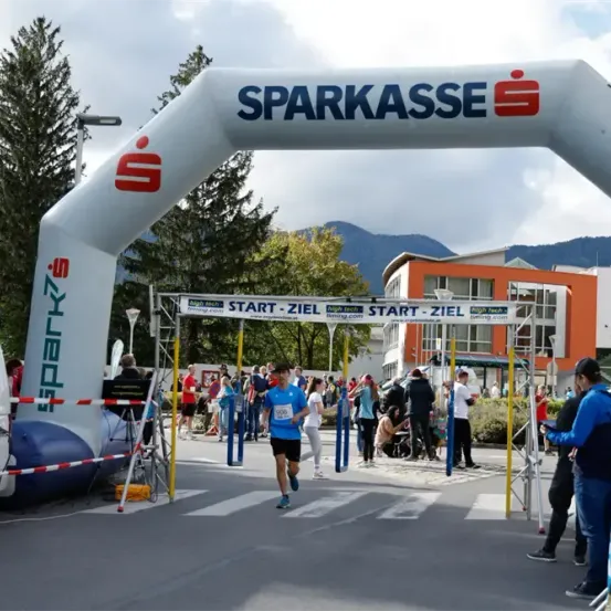 Ein Marathonrennen mit einer Menge Teilnehmer und Zuschauern. Der Torbogen trägt den Schriftzug 'Sparkasse' mit einem Logo. Ein Läufer in einem blauen Trikot läuft durch die Ziellinie. Bäume und Gebäude sind im Hintergrund.