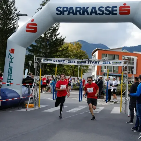 Zwei Läufer in roten Shirts und schwarzen Shorts laufen bei einem Marathonrennen vor einer Menge. Der Wettkampf hat einen Bogen, der 'SPARKASSE' und ein Banner, das 'START-ZIEL' sagt. Der Marathon ist in einer Stadt mit Bergen in der Ferne.