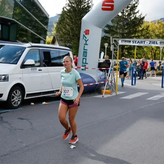 Eine Frau läuft bei einem Marathon in einem grünen Shirt mit der Nummer 307 und passiert einen weißen Bogen mit dem Text 'Start-Ziel'.