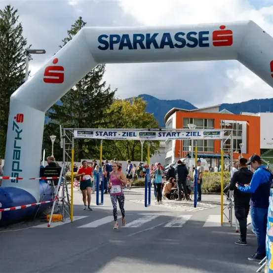 Eine Marathonläuferin durchquert den Zielbogen. Der Bogen wird von 'Sparkasse' gesponsert. Leute gehen herum und schauen sich die Veranstaltung an.
