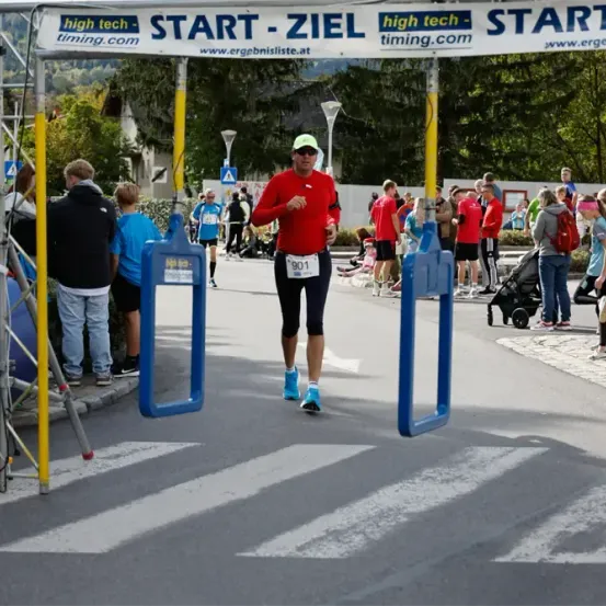 Ein Marathonläufer durchquert eine Zeitnahme-Tor, trägt ein rotes Shirt und blaue Schuhe, während andere am Rennen teilnehmen. Ein Banner zeigt 'high tech timing.com START - ZIEL'.