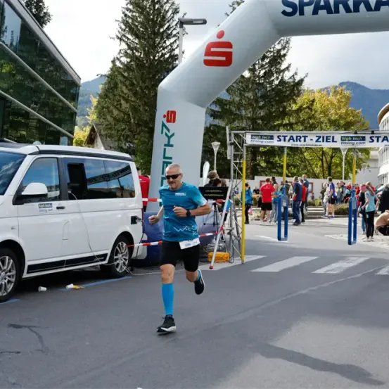 Ein Mann in einem blauen Shirt läuft unter einem weißen Bogen mit der Aufschrift 'Start' und 'Ziel' in einem Außenbereich mit einer Menge von Menschen.
