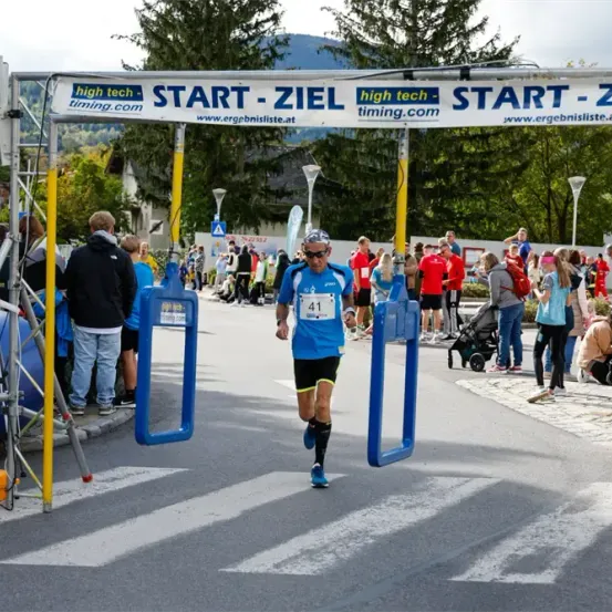 Ein Marathonläufer läuft unter dem Start-Ziel-Banner hindurch, mit Zuschauern und einem Kinderwagen am Straßenrand.